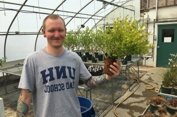 Male student in greenhouse holds a plant. 他的t恤上写着主要研究研究生院.
